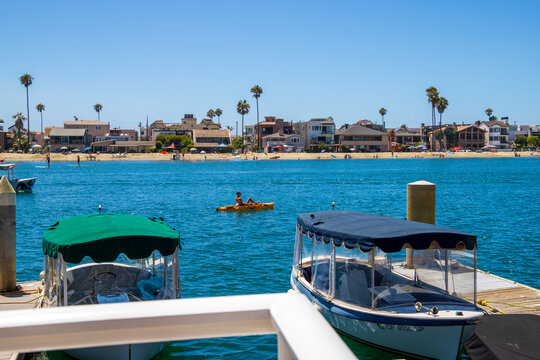 A Gorgeous Summer Landscape At Naples Canals With People Rowing Paddle Boards On Blue Ocean Water With Boats Docked Along The Banks In Front Of Homes With Lush Green Palm Trees And Blue Sky