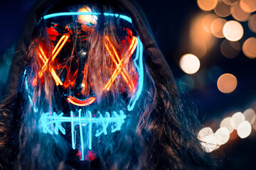 Man in hoodie with long disheveled hair in a lighting neon glowing mask. Halloween dress-up party in night club. Anonymous person is a cyber criminal. All Saints' Night.