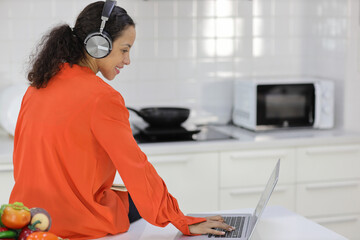 Happy latin woman learn cooking food while checking recipe from internet with computer in kitchen. Young female with headphone prepare delicious meal and listen music. Healthy and lifestyle concept