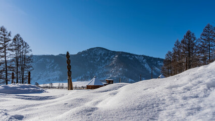 In the snow-covered valley there are traditional Altai houses - ail with a pyramidal roof. Pure white snow in the foreground. A mountain against a clear blue sky. Altai