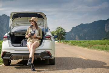 A female traveler with a backpack in a hatchback on a road with mountains, trees and sky in the background.