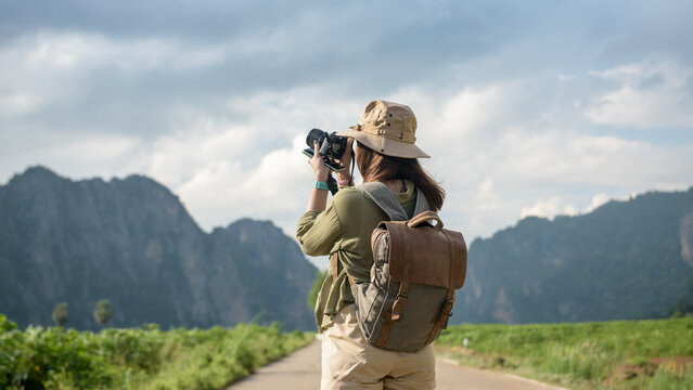 Beautiful young woman backpacking outdoors with analog camera taking pictures on a tourist road with mountains and sky in the background.