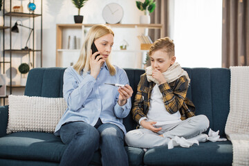 Family, health care and people concept. Portrait of blond woman mother with thermometer and smartphone calling to doctor at home. Sick son sitting on sofa and coughs near his mom.