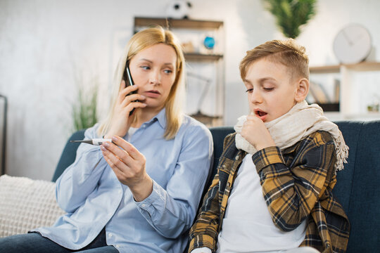Mother holding thermometer and cell phone, calling to doctor, while checking temperature of her sick teen son coughing with high fever sitting on sofa.
