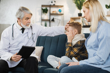 Fototapeta premium Pleasant confident skilled mature man doctor in white coat, visiting patient cute sick preschool boy with his mother, at home, touches forehead to check temperature and giving recommendations.