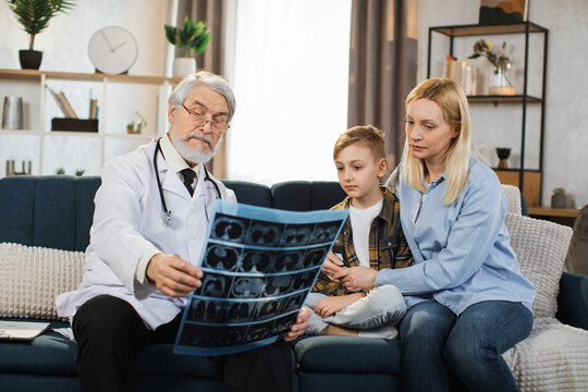 Senior male doctor, sick little boy and his mom looking at x-ray scan image during home visit. Young lady and her child listening their doctor explaining CT scan and talking about the treatment.