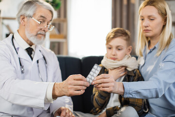 Mature caucasian male doctor giving green blister with tablets for sick preschool coughing boy patient, sitting with his blond middle age mother on the sofa in living room, during home visit.