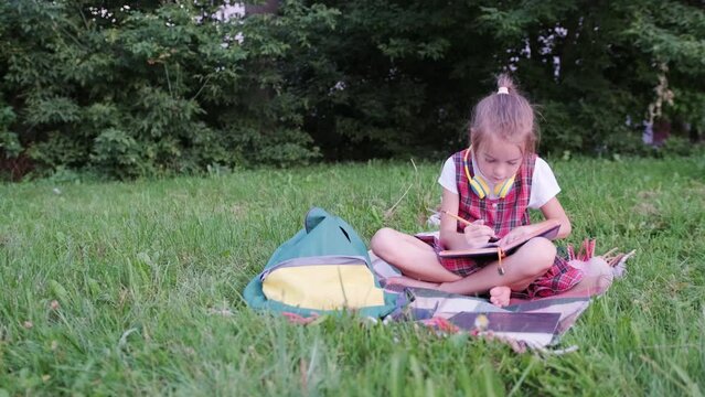 a schoolgirl girl is sitting on the lawn and drawing in a notebook