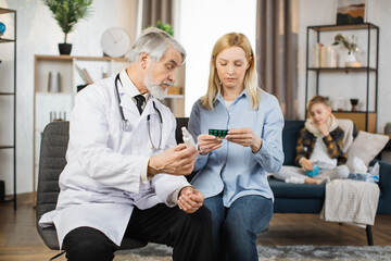 Pretty caring mother sitting with experienced confident mature bearded male doctor and listening...