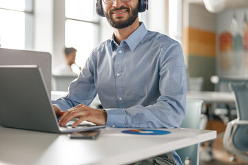 Fototapeta na wymiar Businessman in headphones working laptop while sitting in modern office on colleagues background