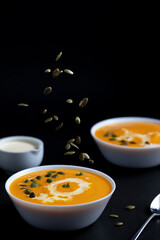 Yellow cream soup in the white dish on the black table and falling seeds