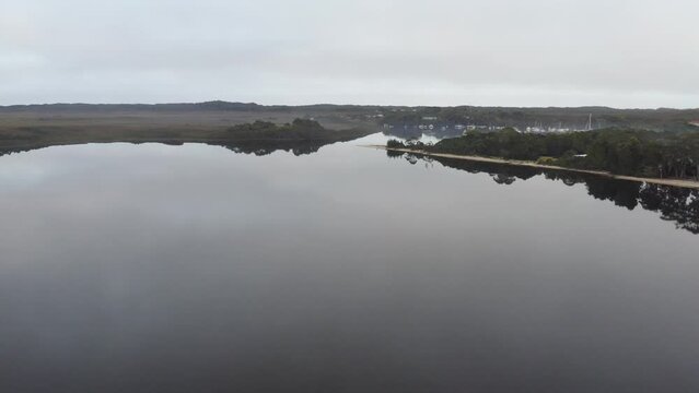 Aerial: Drone flying over a crystal still lake towards a harbour full of boats on a moody morning in Strahan, Tasmania