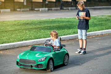 Elder brother controls children car with remote at walk with little brother in public park. Toddler boy sits in green car holding steering wheel looking aside