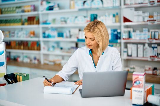 Female pharmacist going through paperwork while working on laptop in pharmacy.