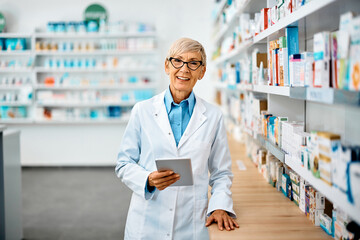 Happy senior pharmacist using digital tablet in pharmacy and looking at camera.