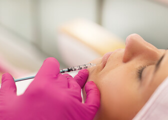 Close-up of young woman getting beauty injection into lips. Cosmetologist making procedure in pink...