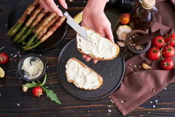 Making breakfast toasts - woman smearing cream cheese on a grilled bread over kitchen table with Asparagus wrapped with bacon and spices on a plate
