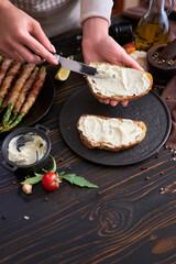 Making breakfast toasts - woman smearing cream cheese on a grilled bread over kitchen table with Asparagus wrapped with bacon and spices on a plate