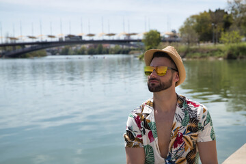 Young and handsome man, mirror sunglasses, beard, hat and Hawaiian shirt, sitting on a pier by the...