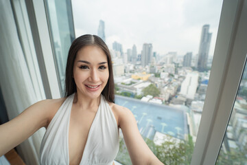 Fototapeta premium A young woman, wearing a white dress, is standing in a room on top of a tall building, behind which is a city view in the heart of Bangkok.