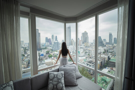 A Young Woman, Wearing A White Dress, Is Standing In A Room On Top Of A Tall Building, Behind Which Is A City View In The Heart Of Bangkok.