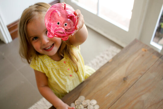Smiling girl with blonde hair wearing yellow blouse raising a pink transparent piggy bank