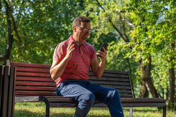 Millennial man in glasses with happy face reading good news on phone while sitting on wooden bench in summer park outdoors. Overjoyed male winner holding smartphone, surprised to receive message