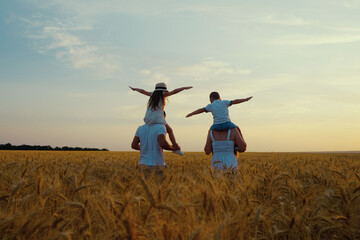 Fototapeta na wymiar Mom and dad carrying kids on shoulders while walking in wheat field at sunset, boy and girl spreading arms as if flying, blue sky on background. Happy family together