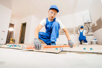 Adult repairman in a special uniform laying tiles with tile leveling system on the floor in a new...