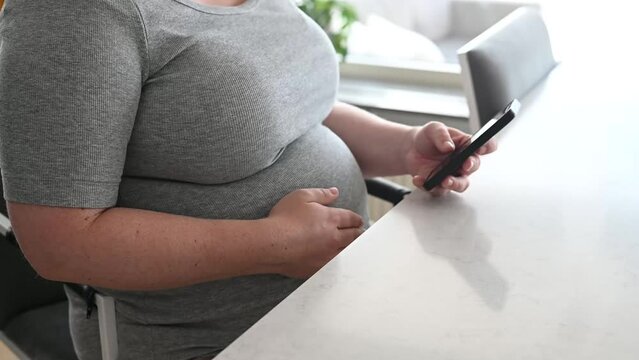 Pregnant woman stroking her belly and using mobile phone at kitchen table at home. Footage made in Sweden.