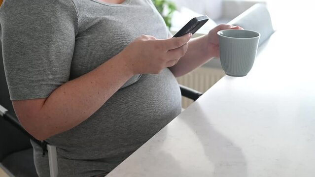 Pregnant woman drinking coffee and using smart phone at breakfast table. Expecting woman in casual clothes sitting at kitchen table at home using mobile phone and relaxing. Footage made in Sweden.