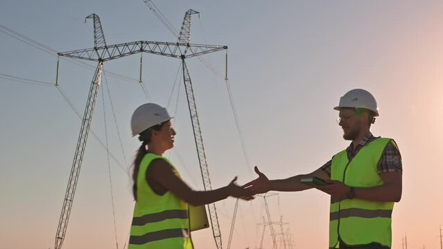 Two electrical engineers meet and shake hands against the backdrop of high-voltage electrical tower at sunset. Meeting of workers near power grids. Teamwork, signing contract, businessmen Slow motion