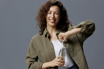 Happy smiling curly beautiful woman in casual khaki green shirt hold open water bottle sticks tongue out posing isolated on gray blue background. Healthy lifestyle. Water is Life concept. Copy space