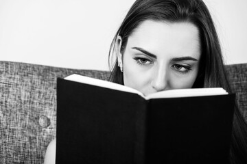 Leisure and hobbies concept. Portrait of beautiful woman with long dark hair reading book. Book with blank black old and aged cover and copy space. Model eyes looking to book. Black and white image