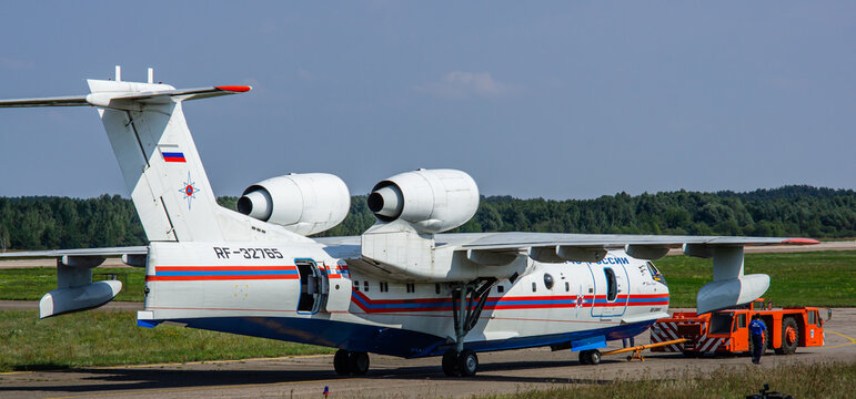 Towing by a tractor of Russian amphibious aircraft (flying boat) - Be-200 "Altair" on taxiway. Close-up. Orange tractor. Blurred background. Selective focus. Zhukovsky, Russia - August 16, 2011