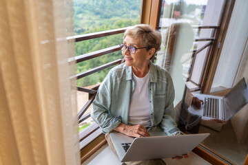 Beautiful happy gray haired senior mature woman in glasses using laptop while sitting by the window...