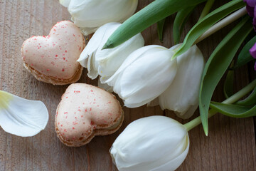 Two creamy macarons are lying on the wooden table with white tulips. Two macarons in form of the heart as 8. Dessert is on the wood