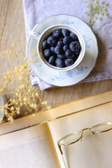Vintage porcelain cup filled with fresh blueberries, pressed flowers, open book, reading glasses and lit candle on the table. Hygge at home. Top view.