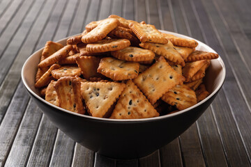 Heap of saltine crackers in a bowl over dark wood plank background. Black deep plate full of salty crackers with black and white sesame seeds. Baked crunchy snack. Saltines concept. Closeup.