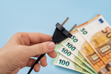 Woman hand holds electric plug against fan shaped euro banknotes over blue background. Energy price...