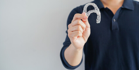 close up young man smiling with hand holding dental aligner retainer (invisible) isolated on gray...