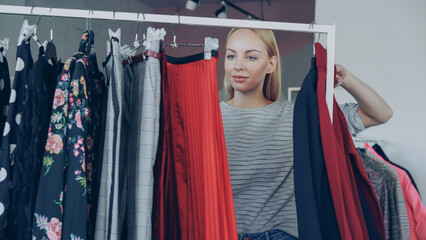 Blonde young lady shopping for clothes, looking through colourful garments on rails, touching and...