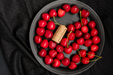 Fresh juicy cherries and wine cork on a black ceramic plate.