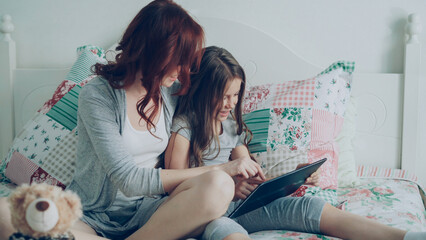 Smiling cute girl and her young mother laughing and using digital tablet while sitting on bed at home in the morning. Technology, home and family concept - Powered by Adobe