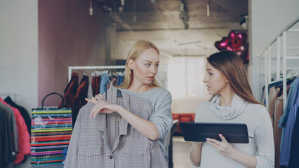Young business owner is using tablet while standing in her clothing store. Her assistant is coming...