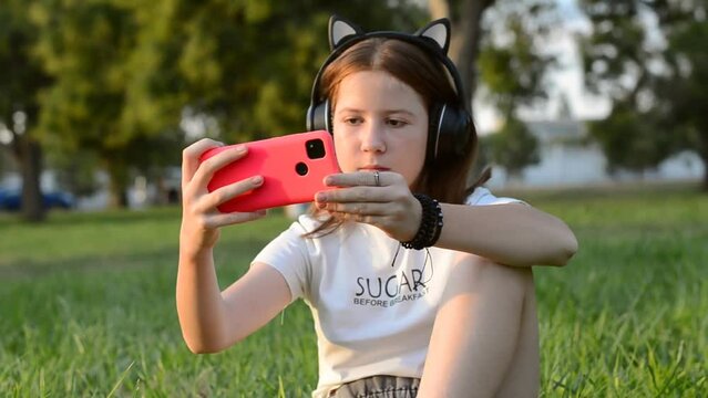 A teenage girl sits in a park on the grass with a smartphone in her hands. 12 year old girl listening to music in headphones sitting in the park on the grass