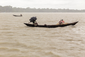Man under an umbrella in a wooden canoe, sheltering from the monsoon rain on a wide river, Barisal,...