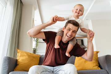 Happy young handsome dad carrying his baby boy on shoulders sitting on sofa posing in front of...