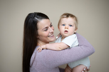 pretty young mother with dark hair, purple top is holding her 7 months old baby and standing in front of brown background and is happy and full of love