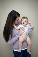 pretty young mother with dark hair, purple top is holding her 7 months old baby and standing in front of brown background and is happy and full of love